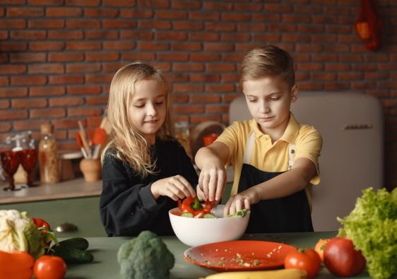Cheerful girl helping little boy in apron preparing salad while standing in kitchen near table with mix of fresh vegetables behind brick wall