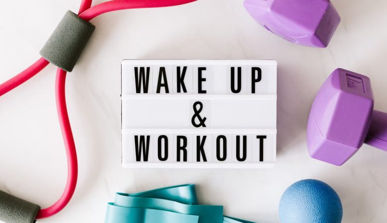 Top view closeup composition of wake up and workout inscription on light box surface among expanders and dumbbells and massage ball on white background